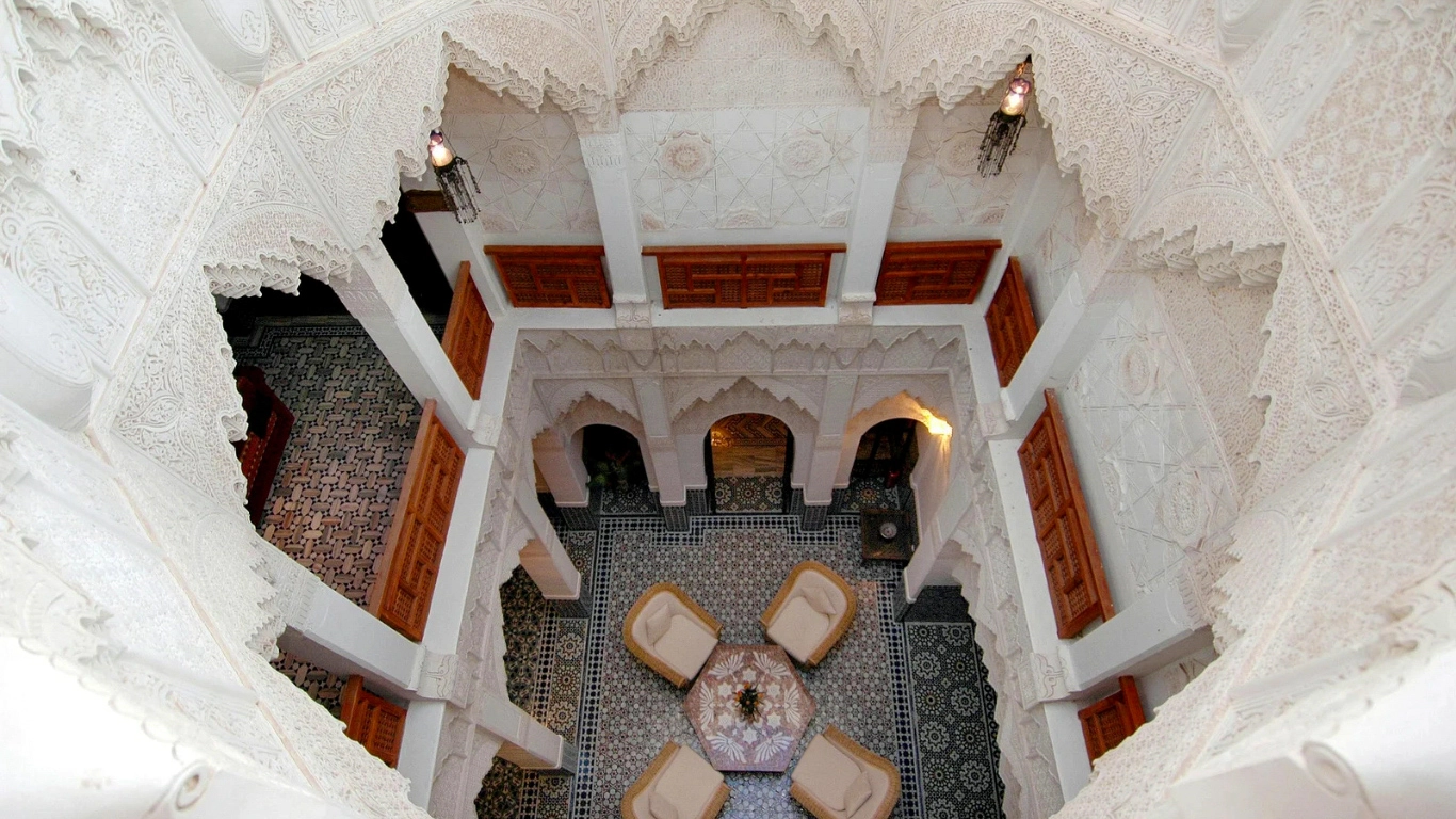 Ornate courtyard of a luxury riad in Marrakech, featuring intricate white plasterwork, traditional Moroccan arches, tiled floors, and elegant seating.
