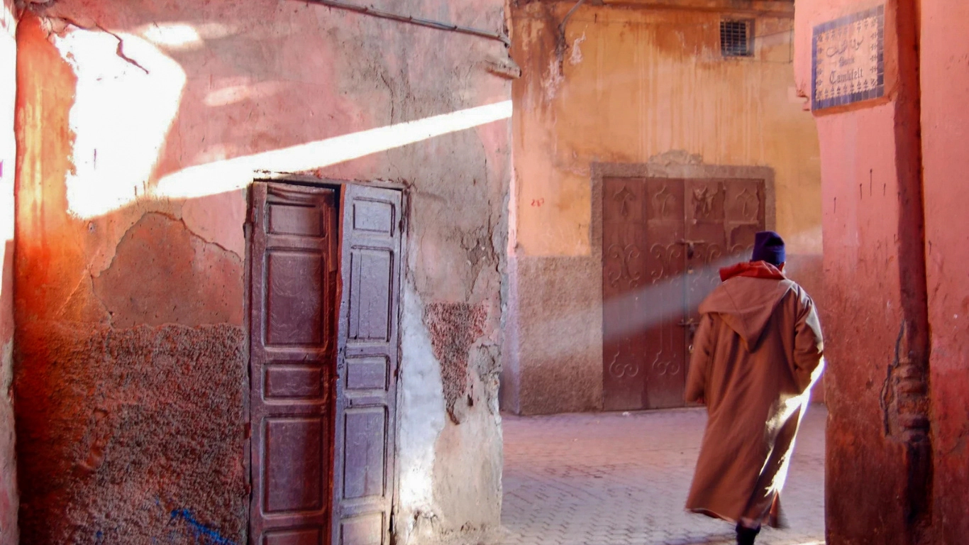 A man in a traditional Moroccan djellaba walks through a sunlit alley with weathered pink and orange walls in the old Medina of Marrakech, capturing the authentic morning atmosphere of the city for a travel article about spending one day in Marrakech.
