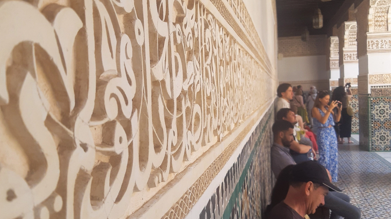 Intricate Arabic calligraphy and zellige tilework at Ben Youssef Madrasa in Marrakech, with visitors admiring the historic architecture during a one-day tour.
