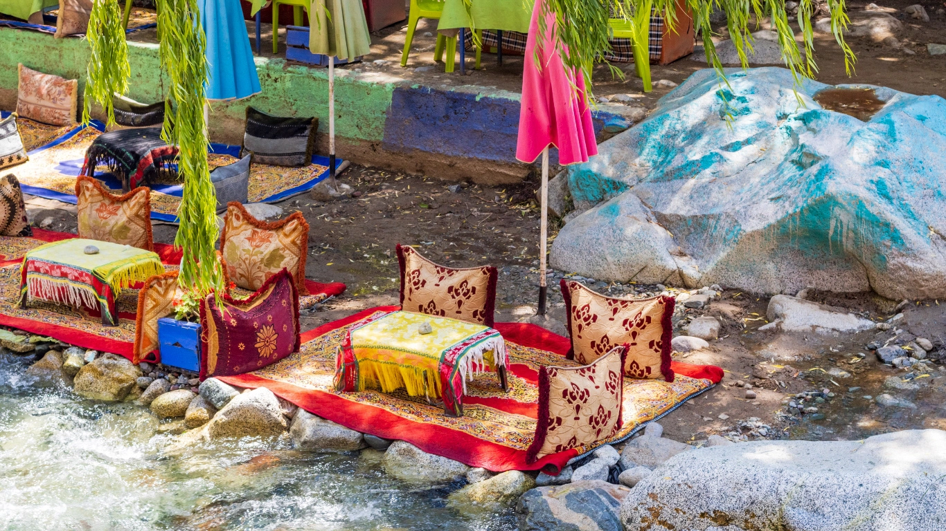 Colorful seating area by a river in the Ourika Valley, surrounded by lush greenery and vibrant decor, ideal for a relaxing day tour from Marrakech.