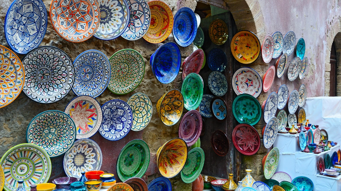 Colorful display of handcrafted Moroccan pottery and ceramic plates hanging on a stone wall in a Marrakech market.