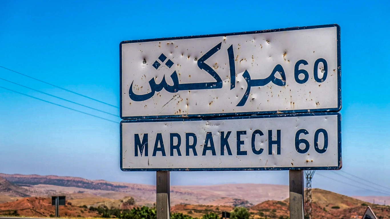 Road sign showing the distance to Marrakech in Arabic and English, with a dry landscape and clear blue sky in the background