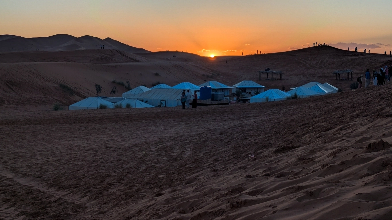 Sunset over the Merzouga desert with Berber tents and sand dunes, showing a peaceful Sahara camp at golden hour.