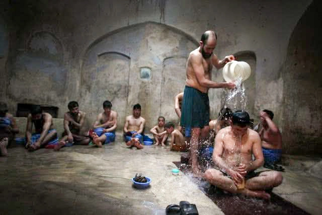 A traditional Moroccan hammam scene showing men bathing and relaxing in a communal bath with tiled walls and wooden buckets
