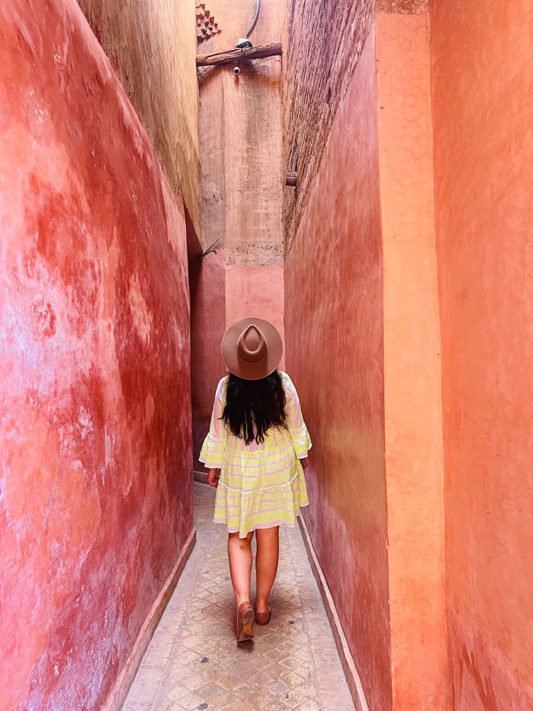 A woman in a yellow dress and hat walks through a narrow, colorful alleyway in Marrakesh, Morocco.